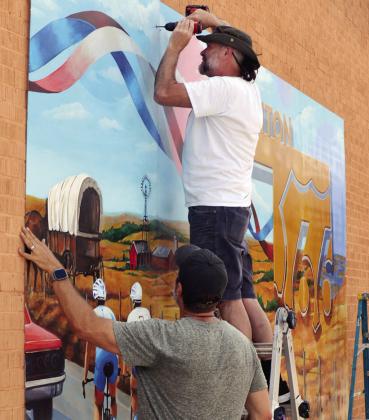 Patrick Hanscom, front, and Scott Henderson install the “American Thread” mural, celebrating the centennial celebration of Route 66 as well as the 250th anniversary of America, on the west wall of S&amp;D Drug. CDN | Christian Jacobsen