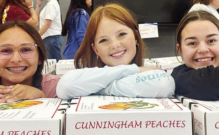 From left, Tatum Fanshier, Bella Bridgeman, and Tatum Shackelford with the Clinton High School Choir stop for a photo as they unload boxes of peaches sold as part of their annual peach sale fundraiser Tuesday afternoon at the high school. CDN | Staff Phot